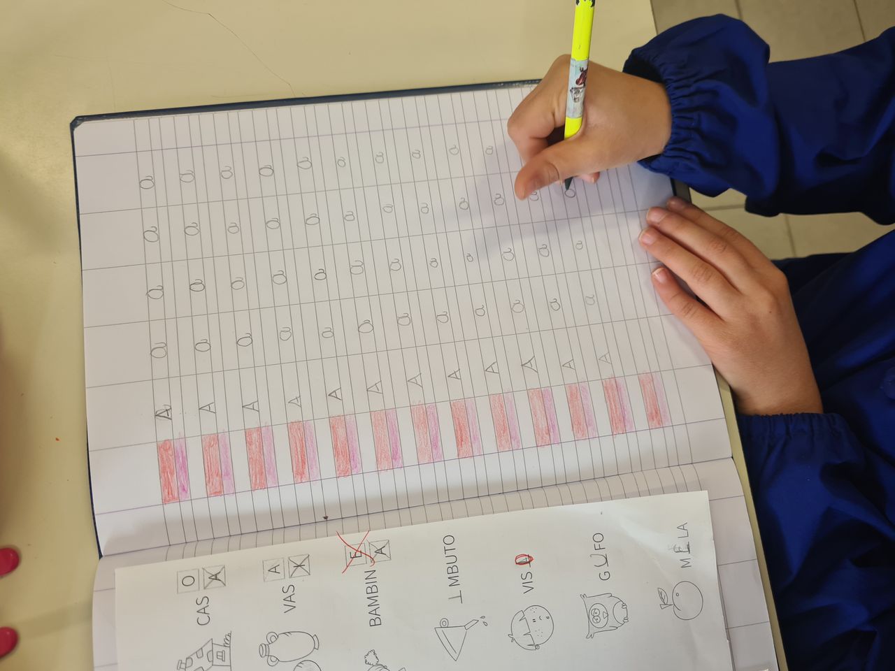 A child in a blue sleeve writes letters in a notebook for educational practice.