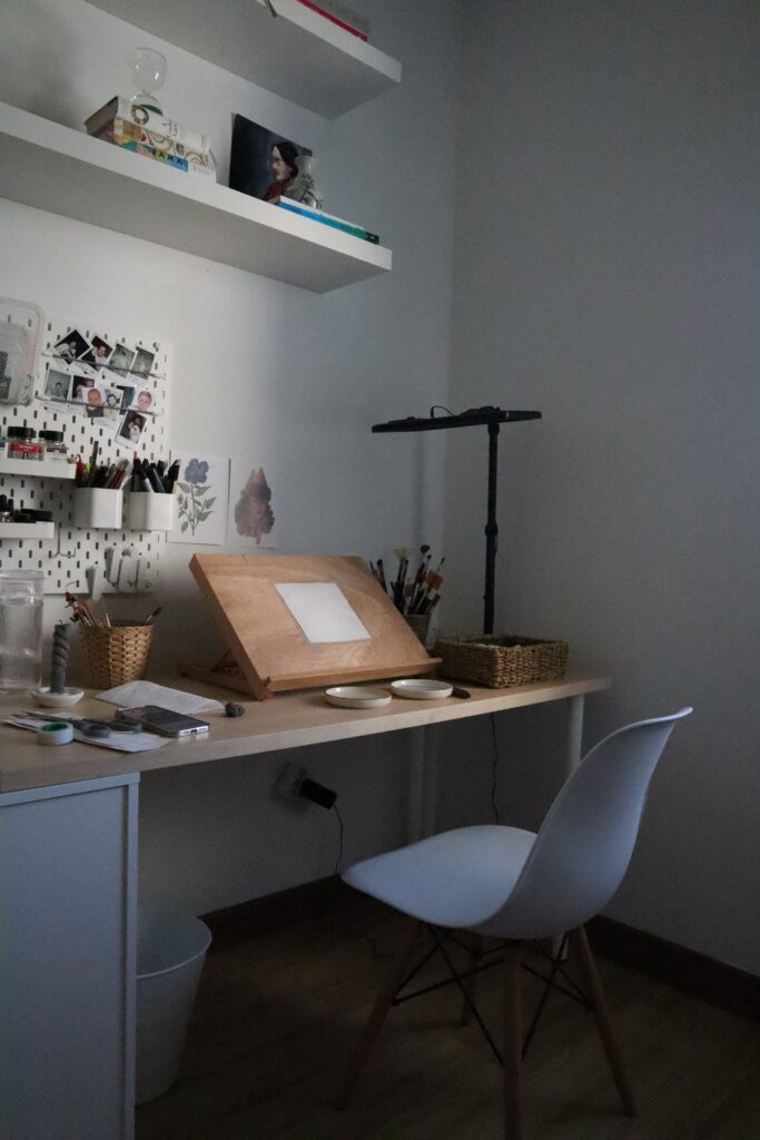 A cozy home office with a desk, chair, shelves, and art supplies.