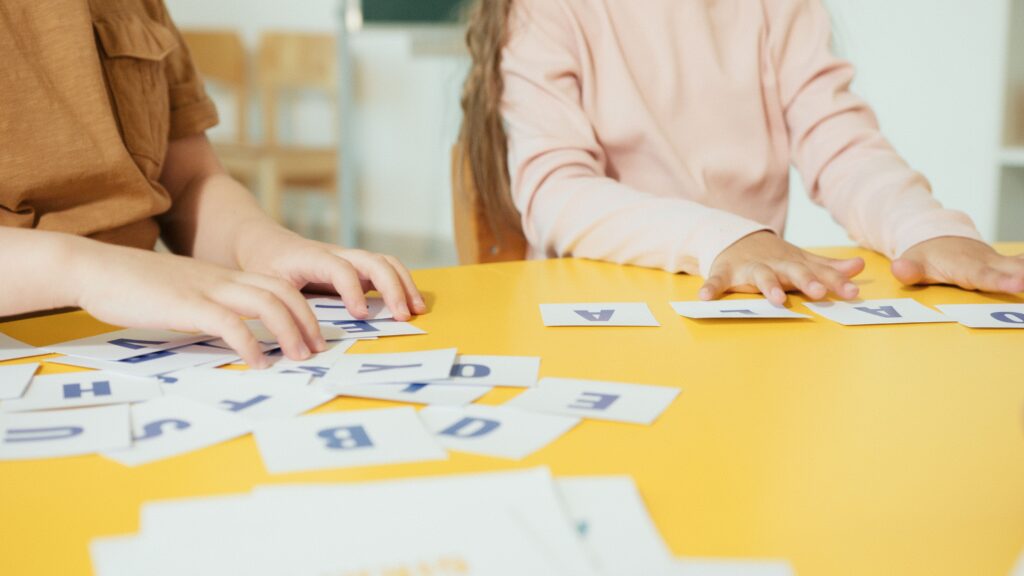 Children arranging alphabet cards on a yellow table in a classroom setting.
